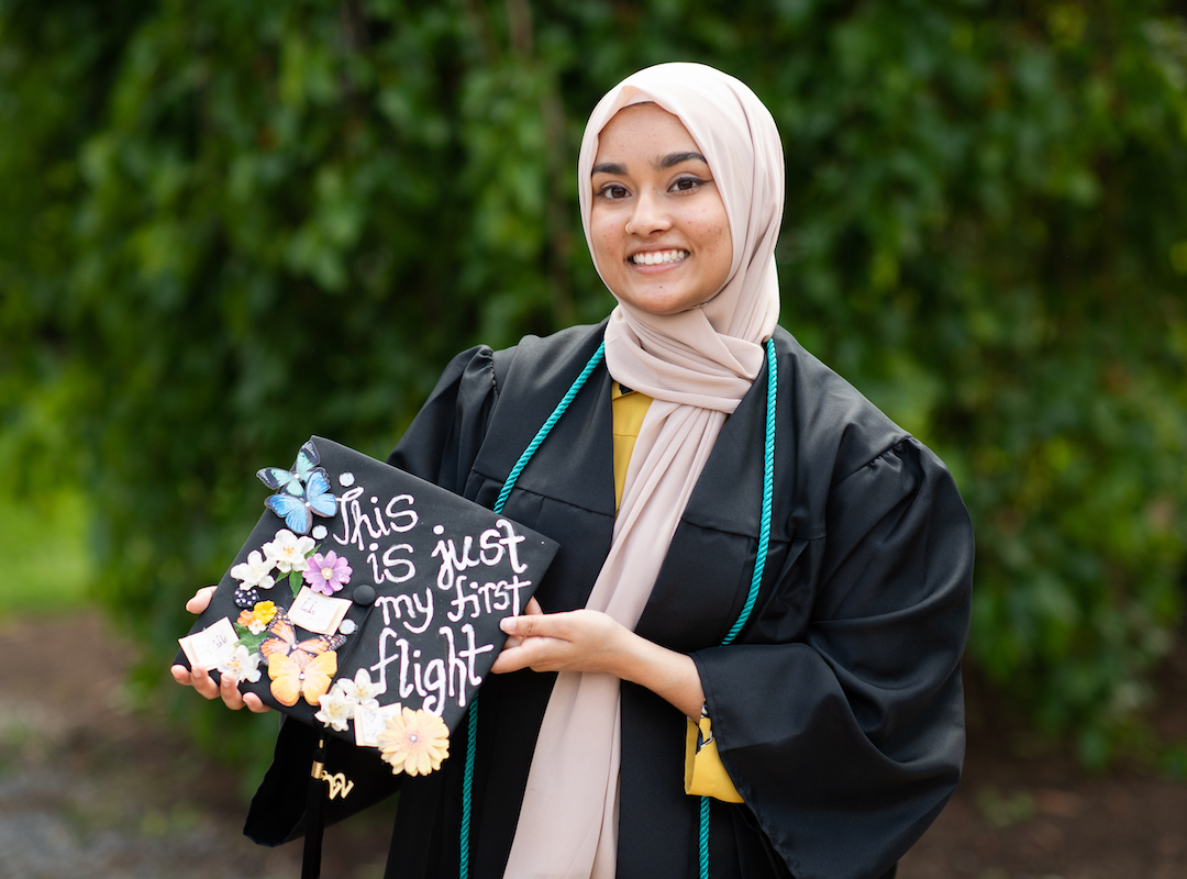Yumna holding a graduation cap decorated with butterflies and flowers and the words "This is just my first flight"