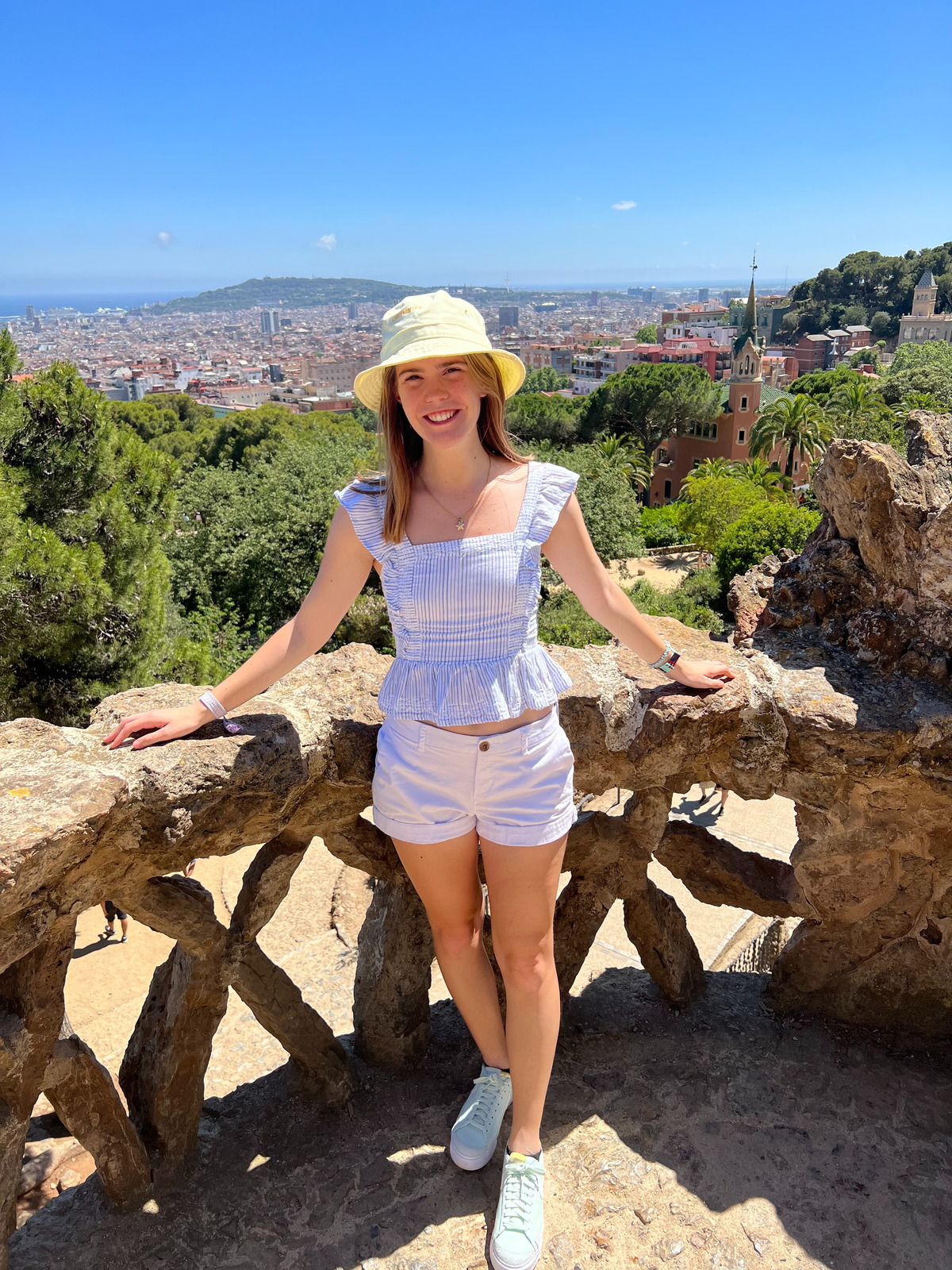 Photo of Aly in a sunhat and shorts and tanktop standing against a railing and blue sky and trees in background