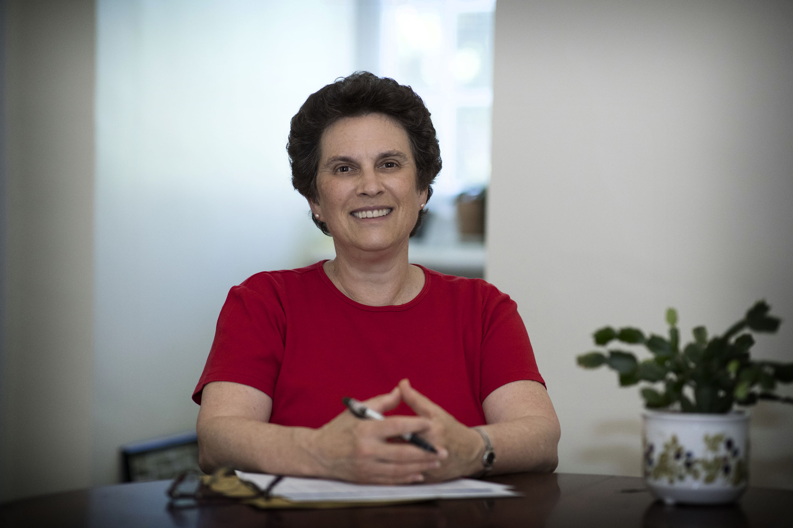 Wynne Stuart sitting at her desk smiling at camera. She is wearing a red t shirt.