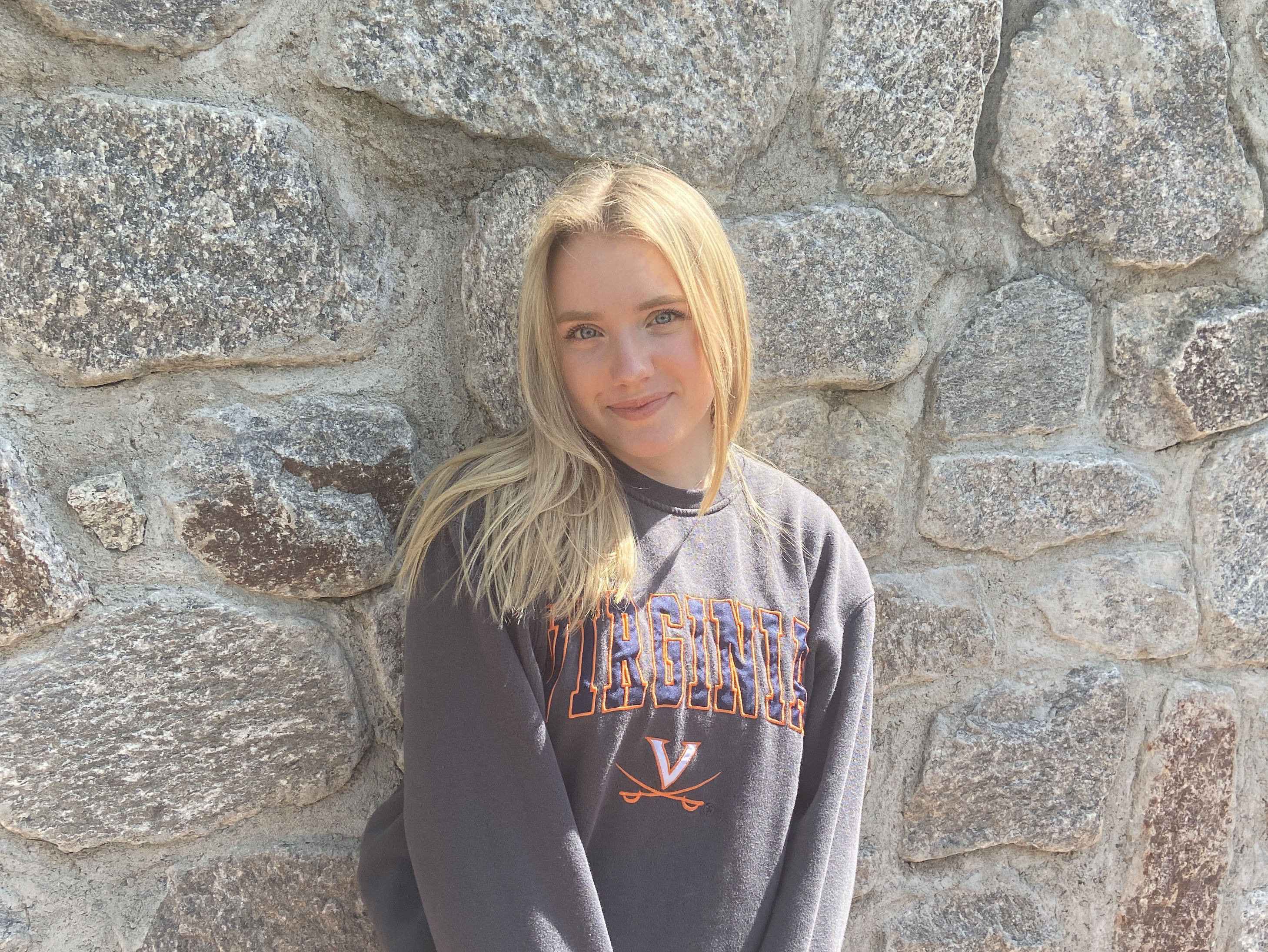 Riley wearing a UVA sweatshirt smiling in front of a cobblestone wall