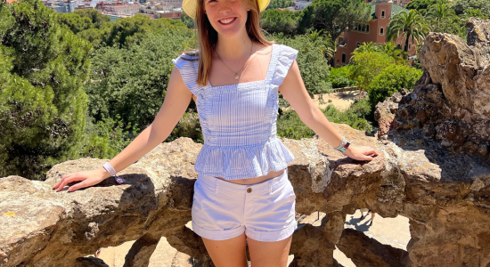 Photo of Aly in a sunhat and shorts and tanktop standing against a railing and blue sky and trees in background