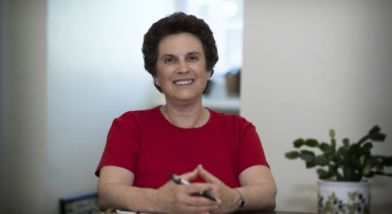 Wynne Stuart sitting at her desk smiling at camera. She is wearing a red t shirt.