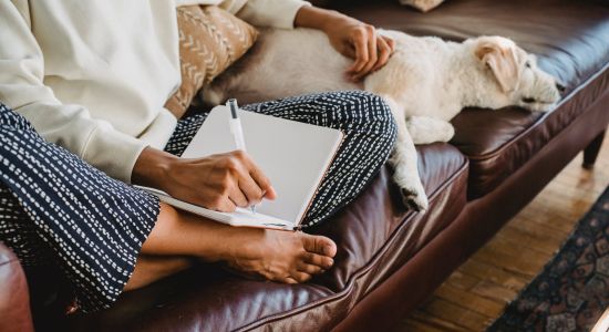 stock photo of someone on couch sitting criss cross with one hand holding pen and writing in journal and other hand on dog laying on couch.